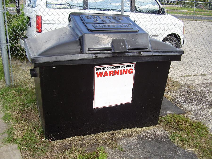 Black outdoor collection bin for spent cooking oil beside a chain-link fence