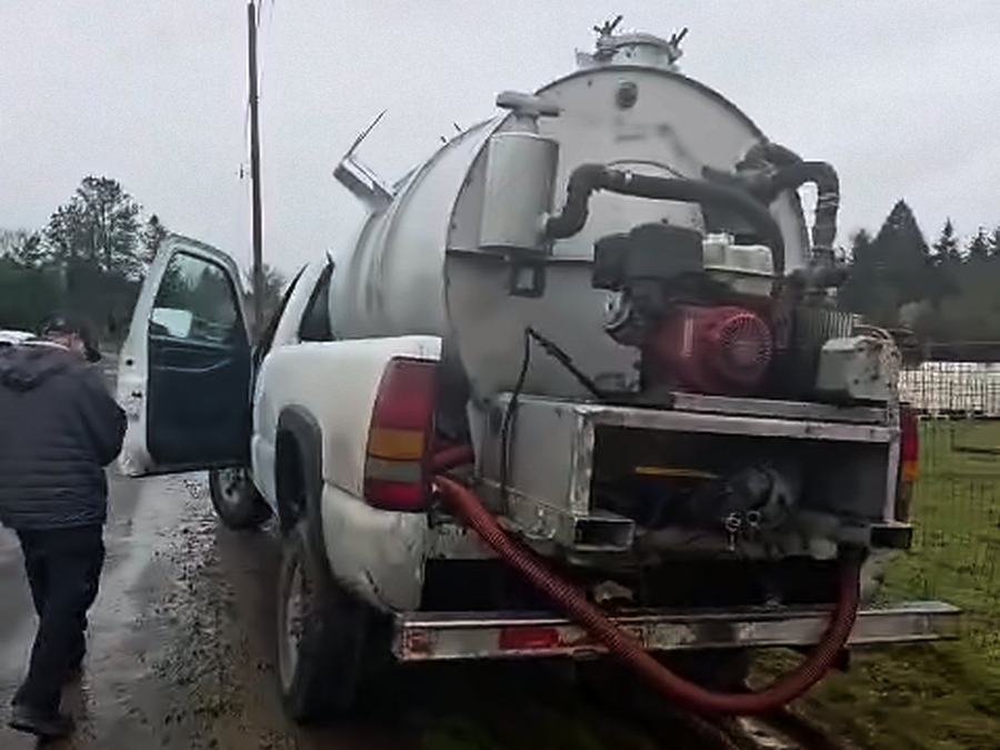Worker walking beside a white pickup with a vacuum tank and pump on the bed, a red suction hose hanging from the back