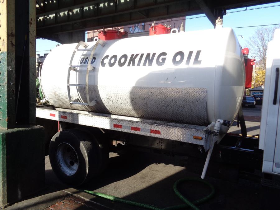 White tank truck lettered Used Cooking Oil at a restaurant stop, with a green suction hose on the pavement