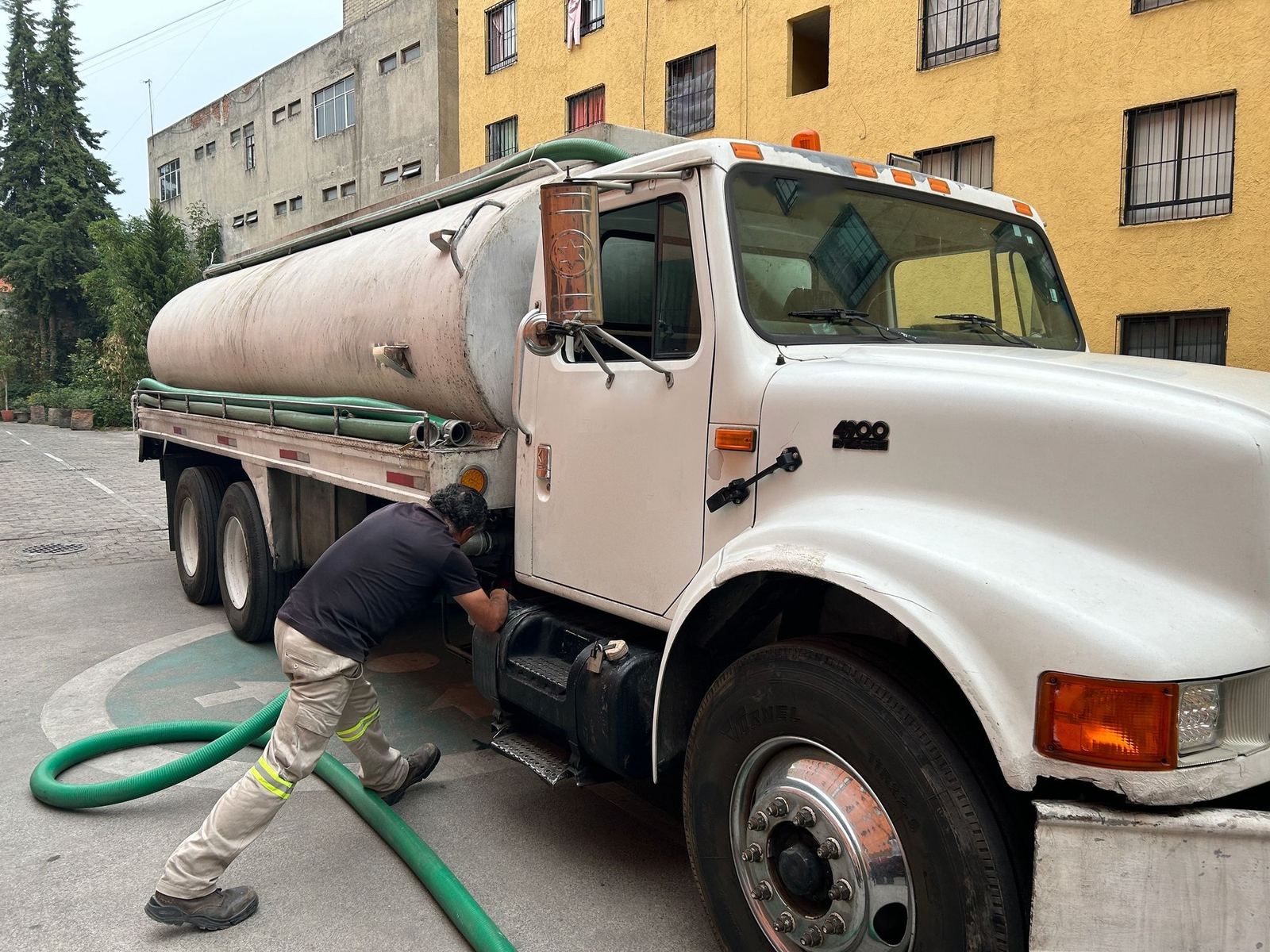 Worker in reflective-striped work pants hooking a green suction hose up to a white tank truck